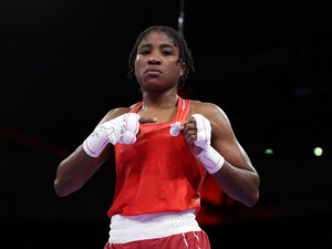 Una mujer posa para una foto con ropa de boxeo color rojo, señalando el logotipo del Equipo Olímpico de Atletas efugiados en su ropa.