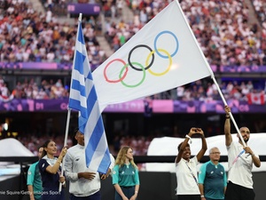 Un hombre porta la bandera olímpica en un estadio abarrotado junto a una mujer que sostiene una bandera griega y otras personas