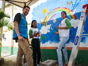 Un hombre con chaleco azul de ACNUR posa con dos niñas adolescentes frente a un muro colorido donde se ve un cielo azul y un arcoiris 