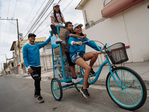 Una mujer pedalea un triciclo y carga a un niño y a una niña atrás. Un hombre los acompaña de pie sobre la calle