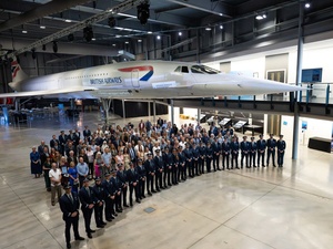 Una fila de pilotos posa delante de un grupo de personas en un hangar con un avión concorde detrás de ellos