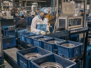 Makendy, un refugiado haitiano en México, trabaja en una línea de producción automotriz en Exedy Dynax, una empresa japonesa en Aguascalientes. Vestido con el uniforme de la empresa y guantes de protección, inspecciona cuidadosamente un componente, rodeado de maquinaria y bandejas organizadas con piezas. Tras participar en el Programa de Integración Local, obtuvo un empleo formal, aportando sus habilidades y experiencia a la industria.
