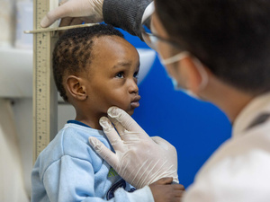 Un niño con sudadera azul claro frente a una regla mientras un hombre con mascarilla y guantes mide su estatura