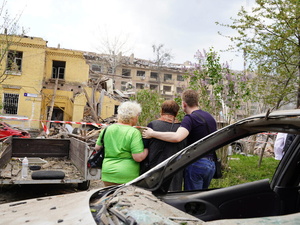 Tres personas fotografiadas de espaldas miran hacia unos edificios gravemente dañados y rodeados de escombros.