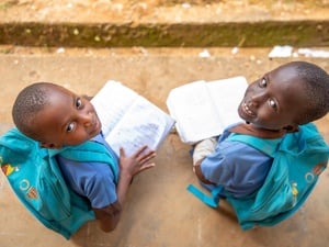Dos niñas de 7 y 8 años, procedentes de la República Centroafricana, sonríen a la cámara desde donde están sentadas al aire libre, con mochilas a juego en las que se lee 'La educación no puede esperar' y sosteniendo libros abiertos