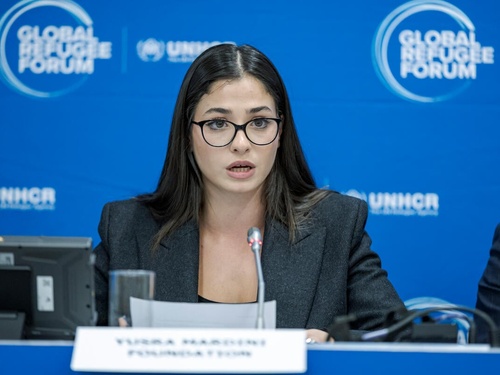 Una mujer joven, que usa anteojos, pronuncia unas palabras frente a un micrófono; detrás suyo, un fondo azul con el logotipo del Foro Mundial sobre los Refugiados.