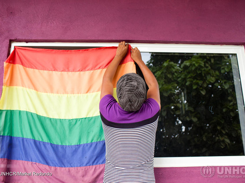 Una persona está colgando una bandera LGBTIQ+ en una ventana; no se puede ver su cara porque le está dando la espalda a la cámara