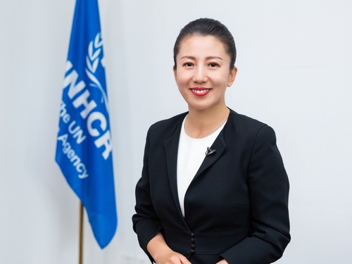 Una mujer china, que lleva puesto un traje color negro, sonríe a la cámara; detrás suyo, una bandera azul con el logotipo de ACNUR en color blanco.