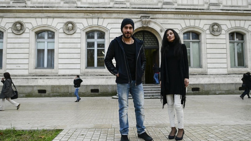 Un hombre y una mujer jóvenes en una plaza viendo a la cámara; detrás suyo, un edificio blanco