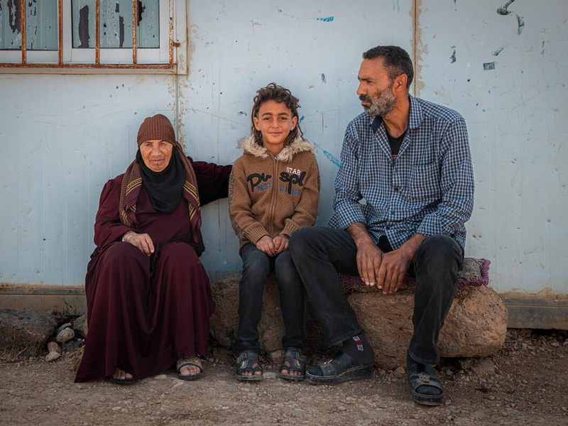 Una mujer mayor de Siria está sentada, junto con su hijo y su nieto, en una banca en el campamento de refugiados de Za’atari.