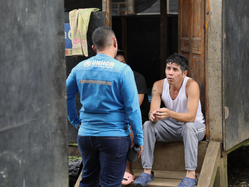 Sentado frente a una puerta de madera, un hombre joven charla con otro que está de pie delante suyo