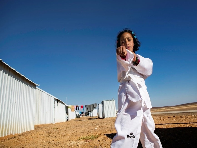 En un campamento de refugiados, una niña que lleva puesto un uniforme blanco de karateka hace un movimiento de karate frente a la cámara