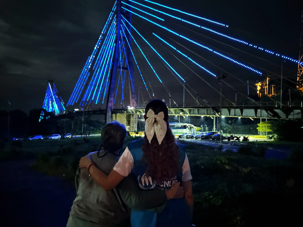 Dos mujeres se abrazan frente a un puente iluminado de azul