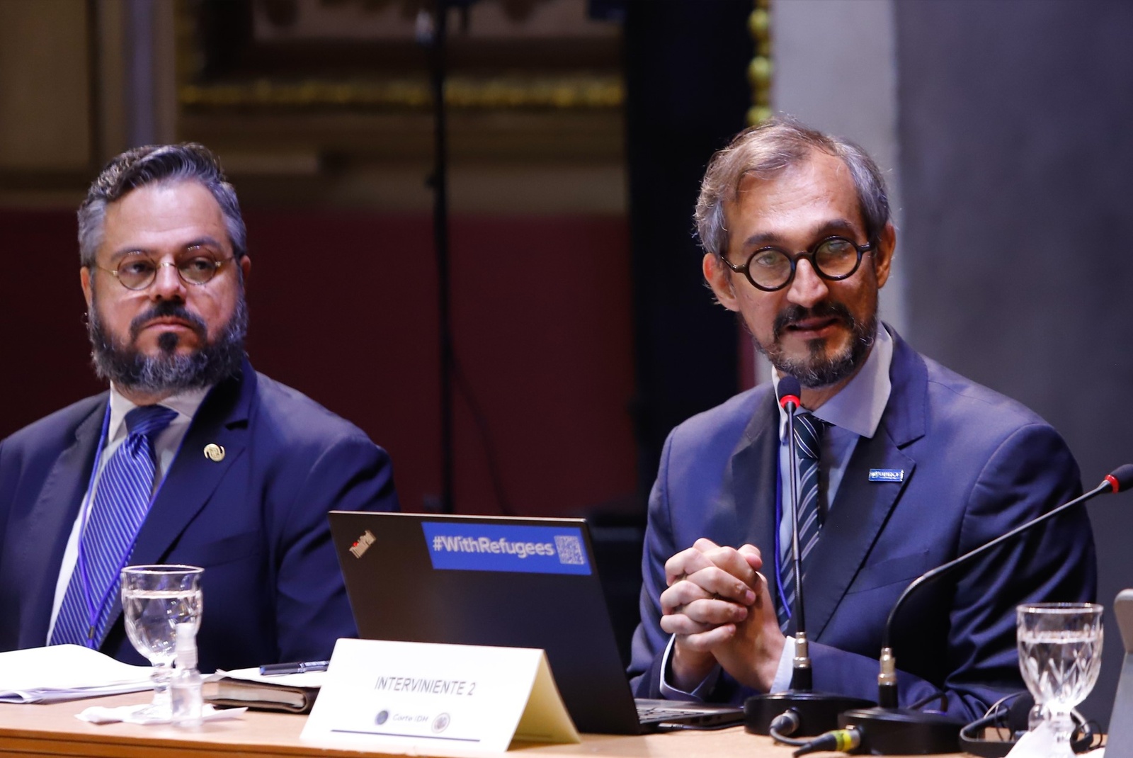 dos hombres de traje oscuro asistiendo a una sesi&oacute;n oficial, uno de ellos est&aacute; hablando.