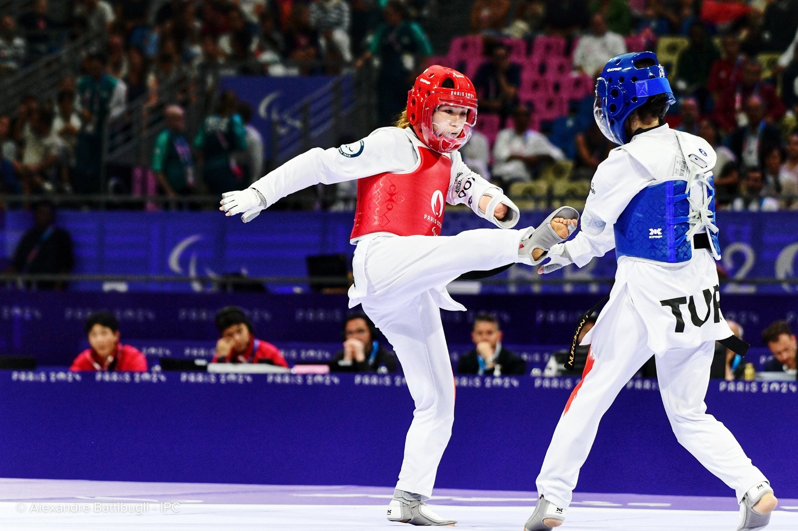 Una atleta de taekwondo con protecciones rojas lanza una patada a su oponente de azul