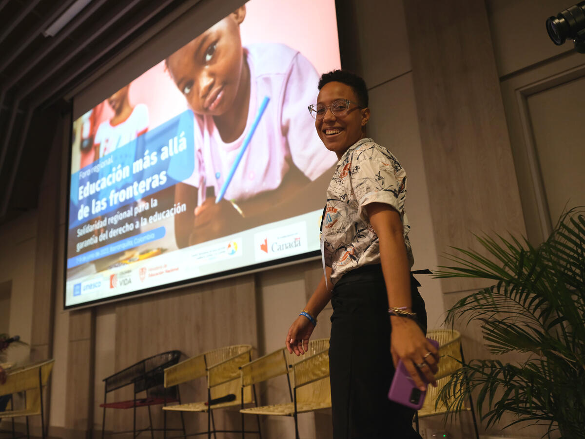 En contrapicada, una mujer de camisa blanca con colores y pantalón negro sonríe para la foto durante una conferencia 