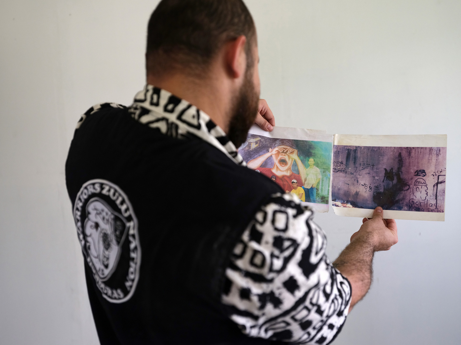 De espaldas, un hombre con camisa blanco con negro y chaleco negro muestra unas fotografías