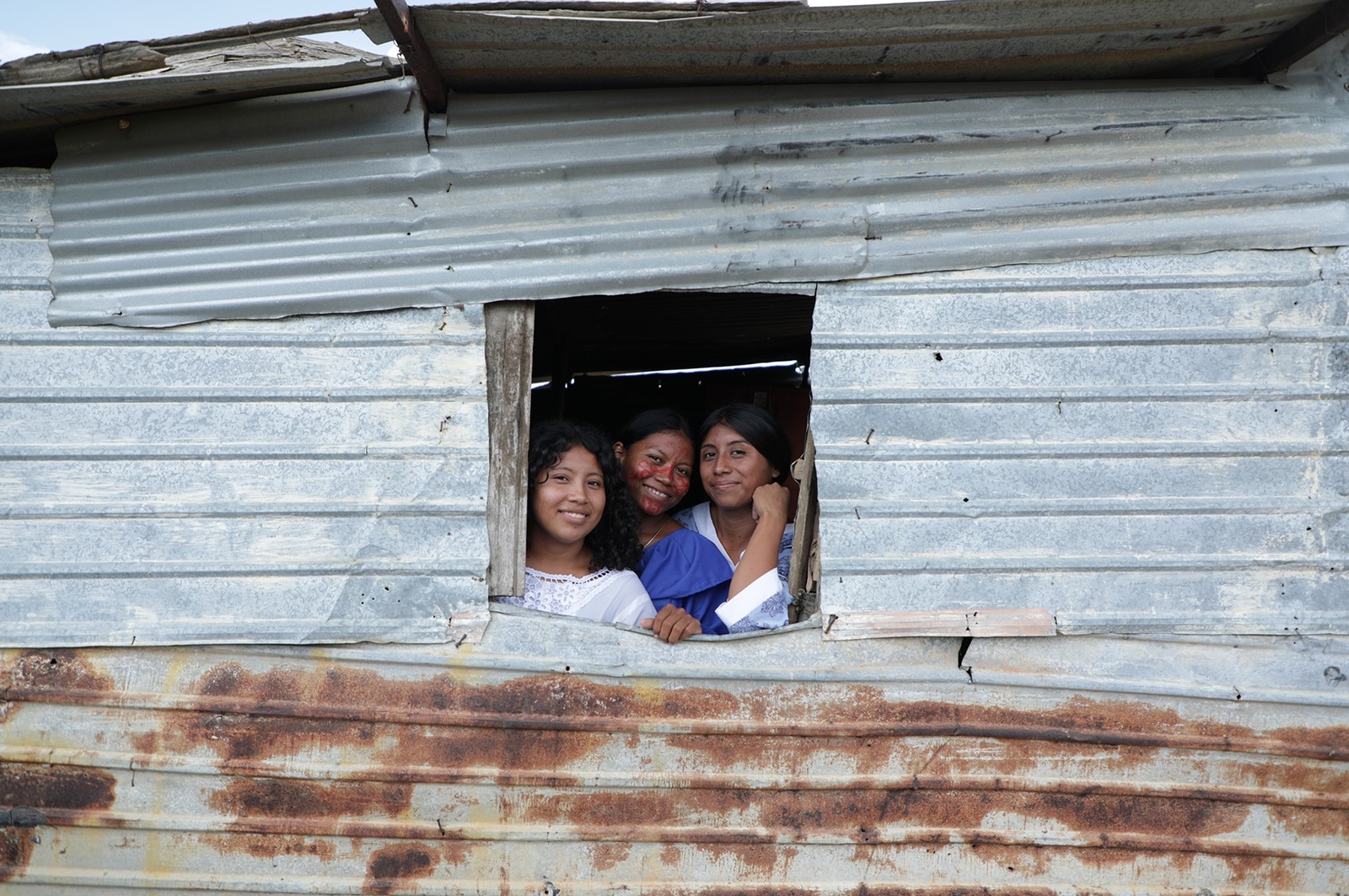 Tres mujeres se asuman a una ventana de una casa con pared de zinc.