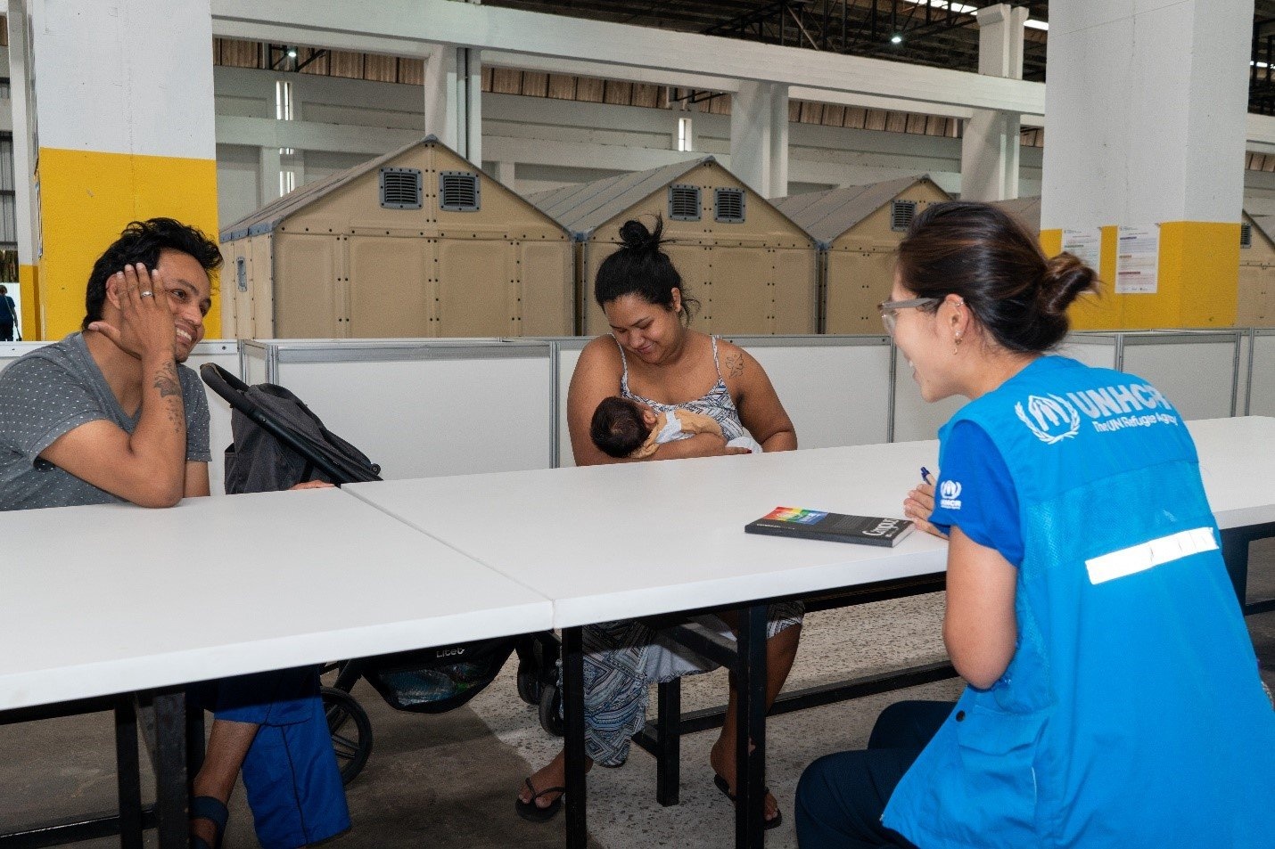 Tres personas hablan sentadas a una mesa, una de ellas lleva un chaleco azul con el logo de ACNUR.