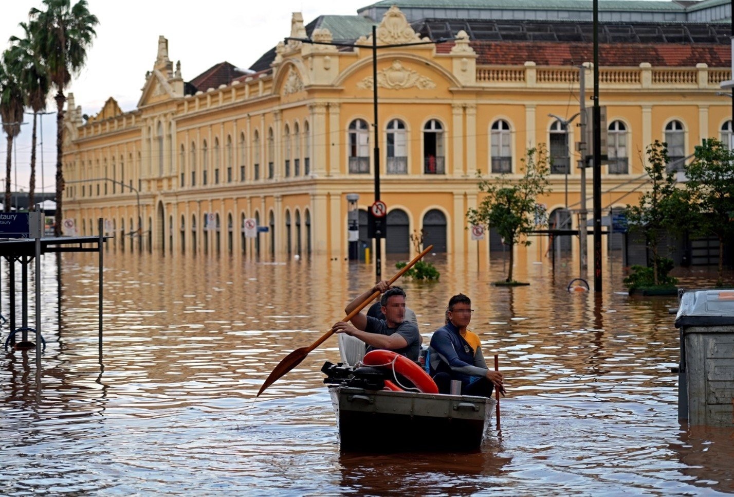 Dos personas se desplazan en un bote en una calle complemtamente inundada en un centro urbano. A sus espaldas un edificio en estilo clásico.