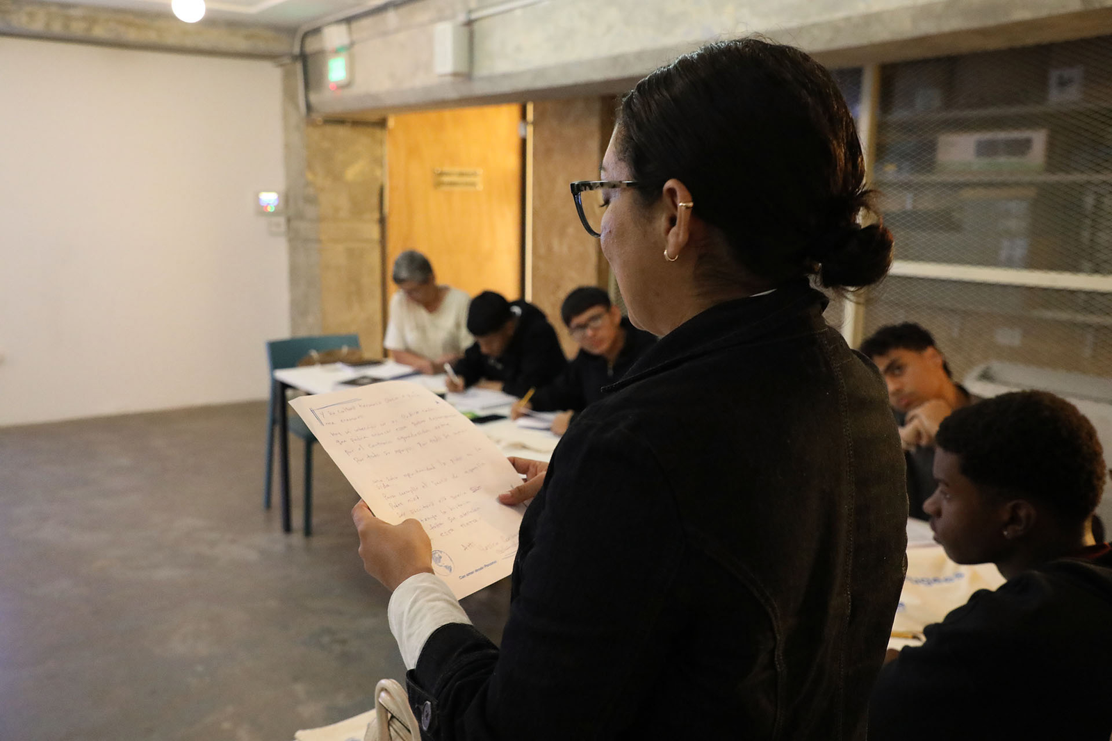 Una mujer fotografiada de espaldas hablando en un salón de clases.