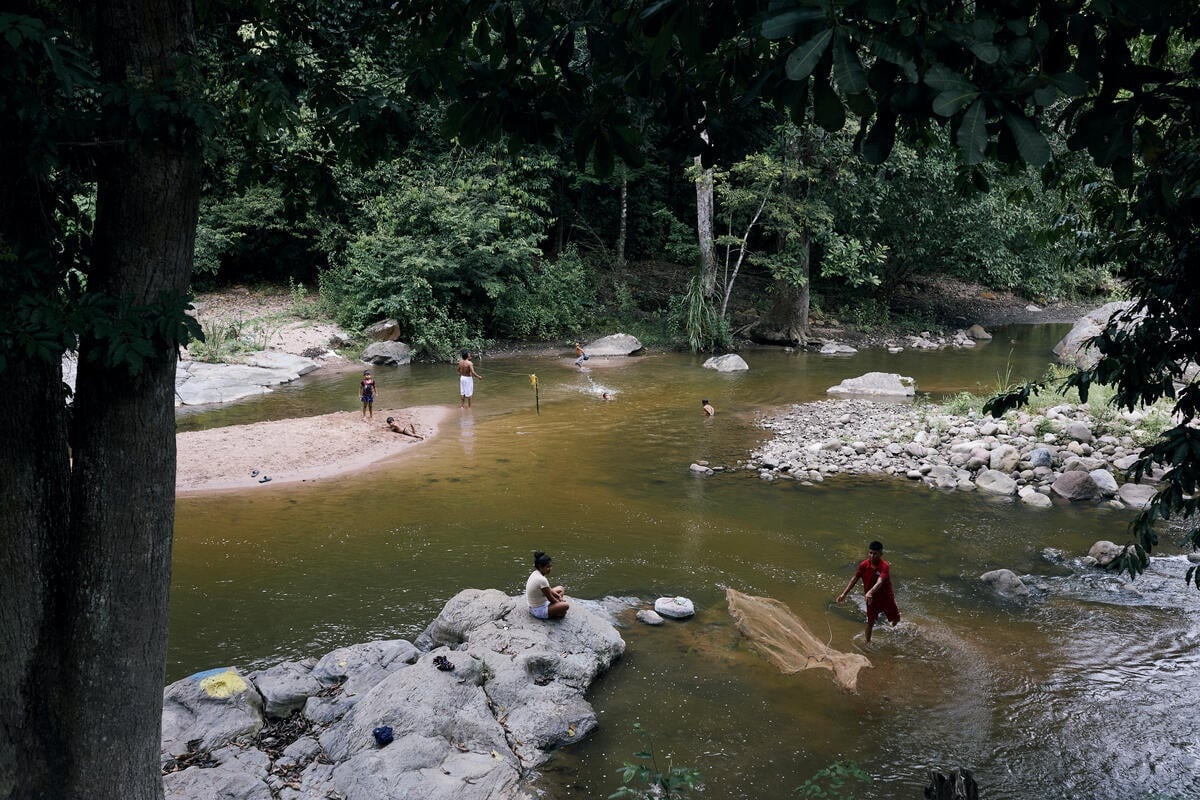 Una vista aérea de un río muestra a un hombre lanzando una red de pesca y a unos niños nadando cerca mientras otros miran desde la orilla
