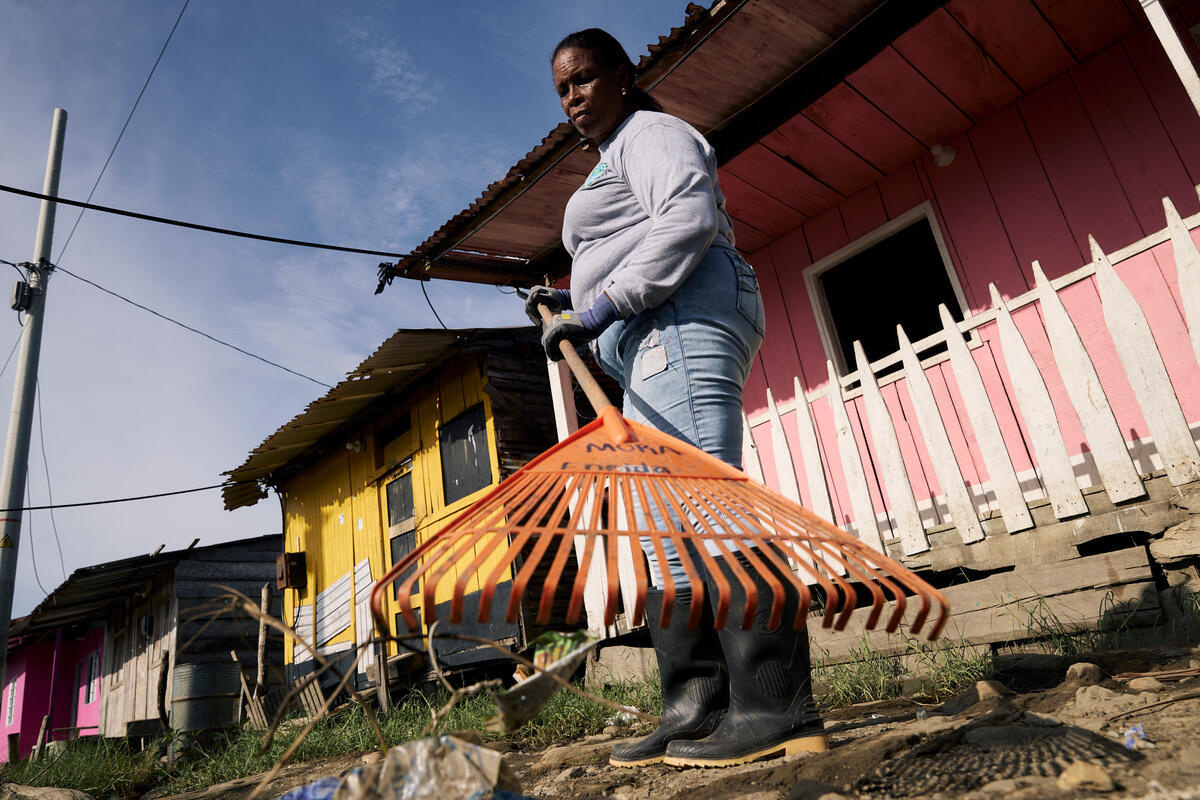 Una mujer afrocolombiana sostiene un rastrillo para recoger desechos; destrás suyo, un par de casas con fachadas coloridas.
