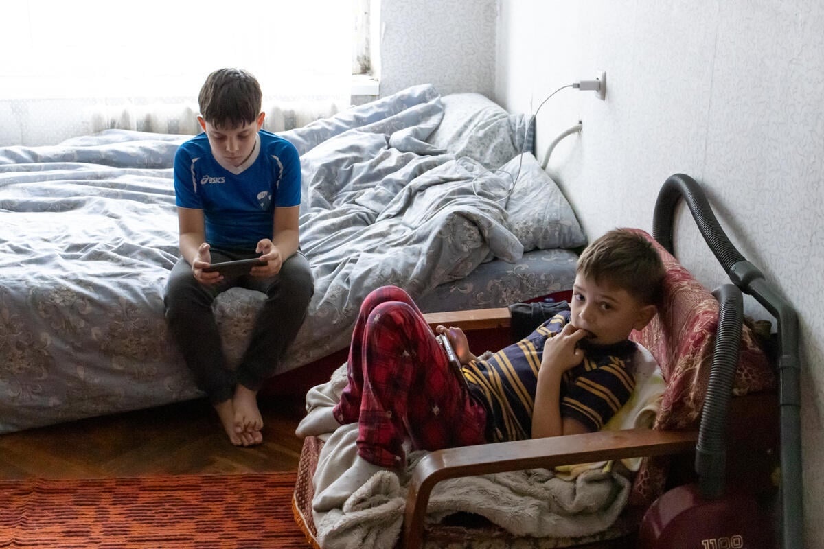 Dos niños juegan con sus teléfonos en su apartamento. Uno está sentado en una silla, con los dedos en la boca, mirando a la cámara, mientras el otro está sentado en la cama