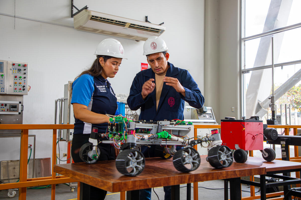 Yuliveth y un técnico de laboratorio del departamento, equipados con casco y ropa de trabajo, comprueban el funcionamiento de un robot con ruedas en el laboratorio.