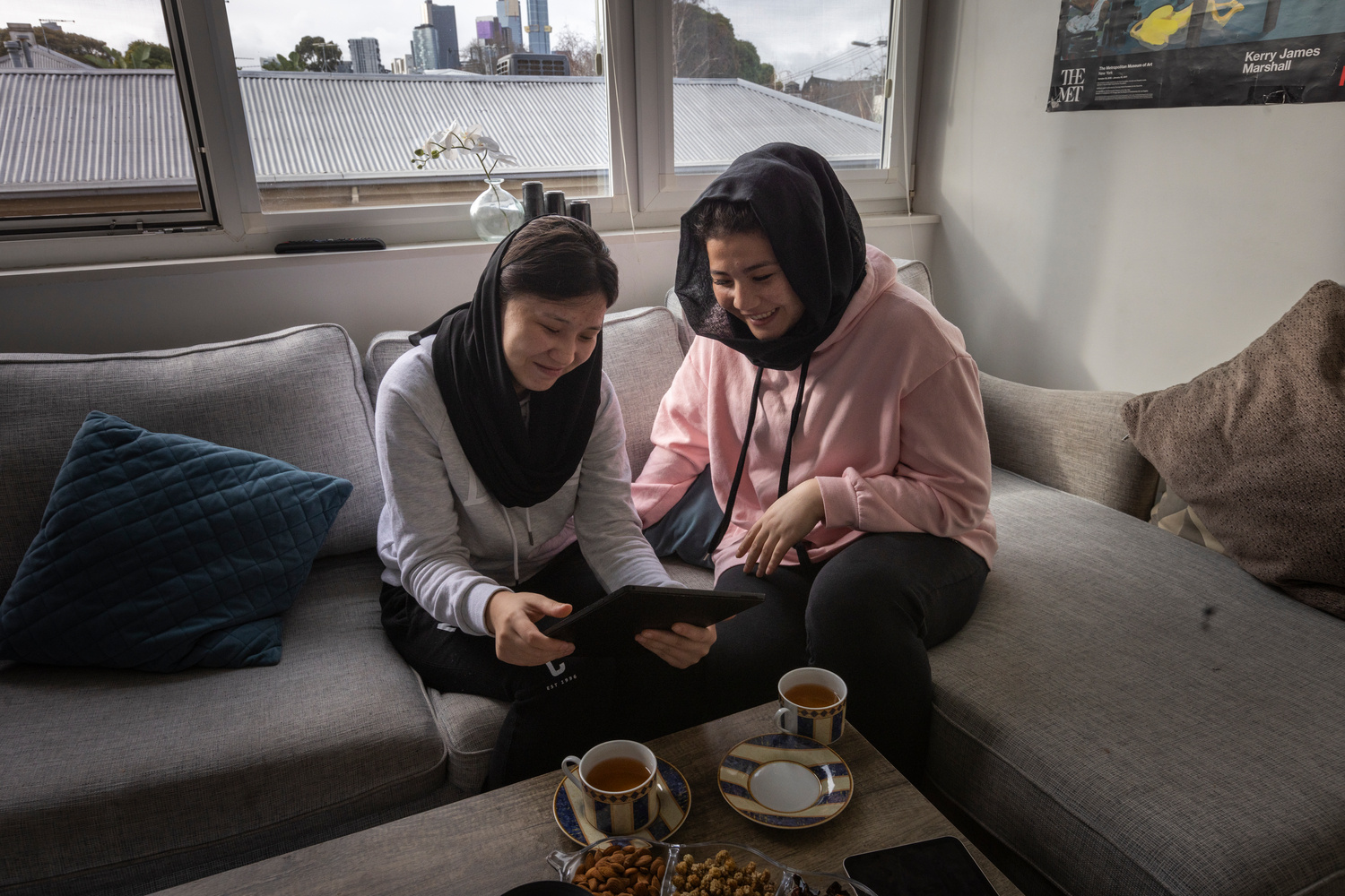Dos jóvenes están sentadas en un sillón, una junto a la otra, observan un tableta y toman té.