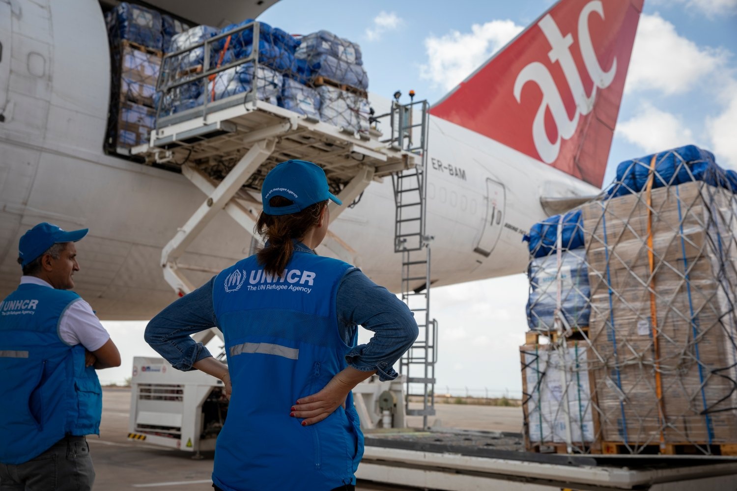 Un hombre y una mujer que llevan puestos chalecos con el logo de ACNUR observan cómo se suben suministros a una aeronave