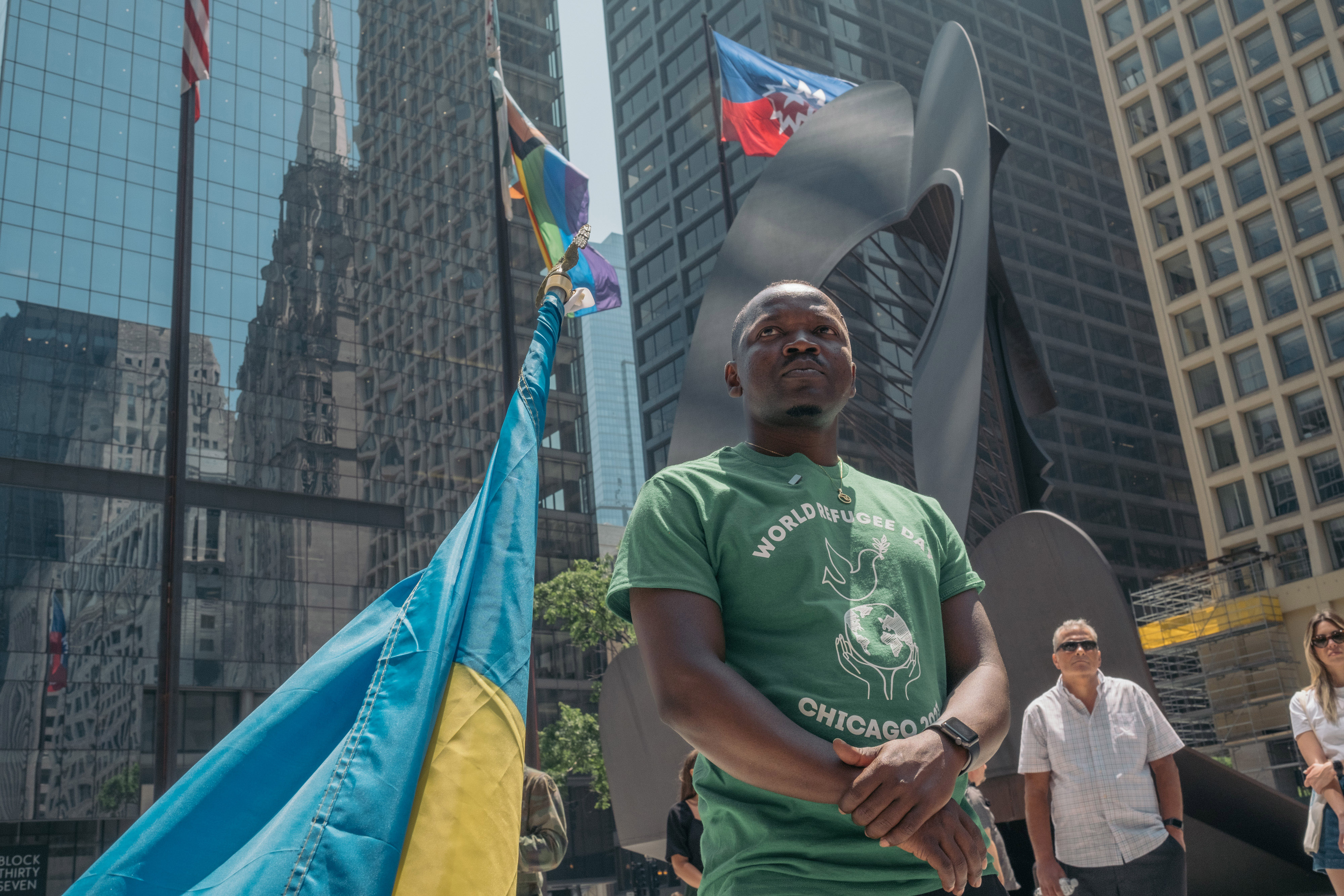 Un hombre está de pie frente a un edificio; con los brazos cruzados ve al horizonte. A lado suyo hay una bandera color azul con amarillo.