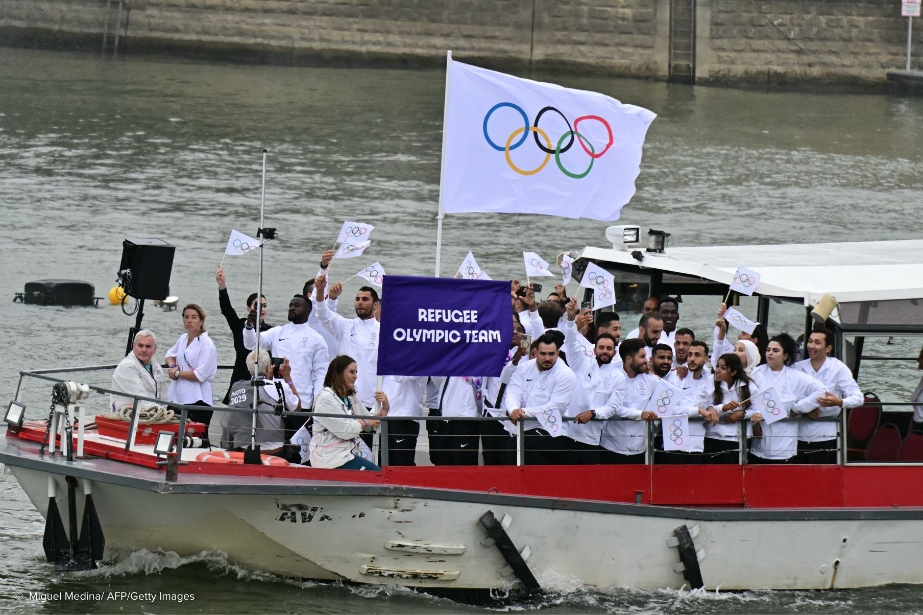 Un grupo de personas vestidas de blanco ondean banderas que muestran los anillos olímpicos en un barco que transita por un río.