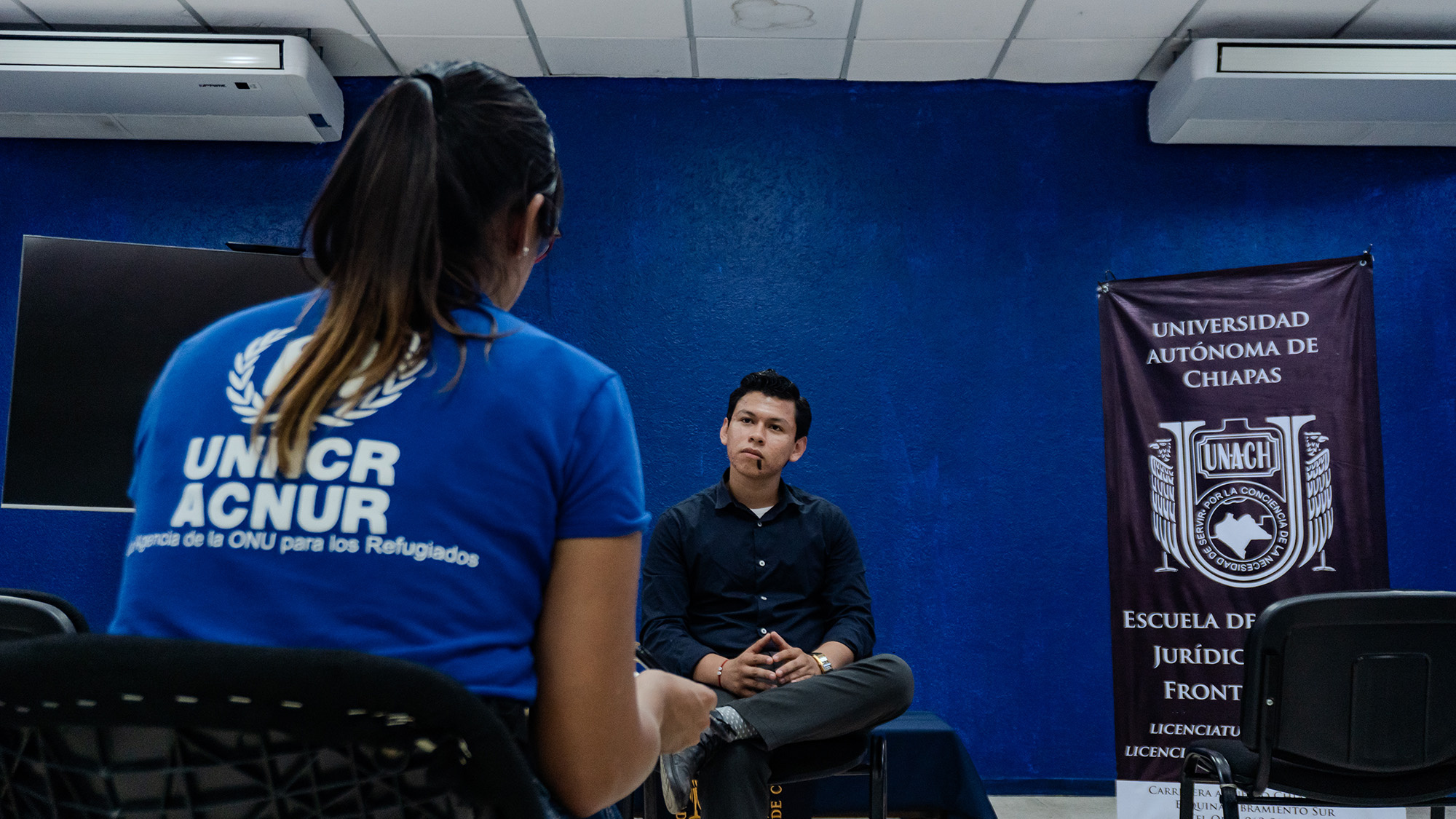 De espaldas, una muejr de playera azul con logo blanco de ACNUR, habla con un joven vestido de negro