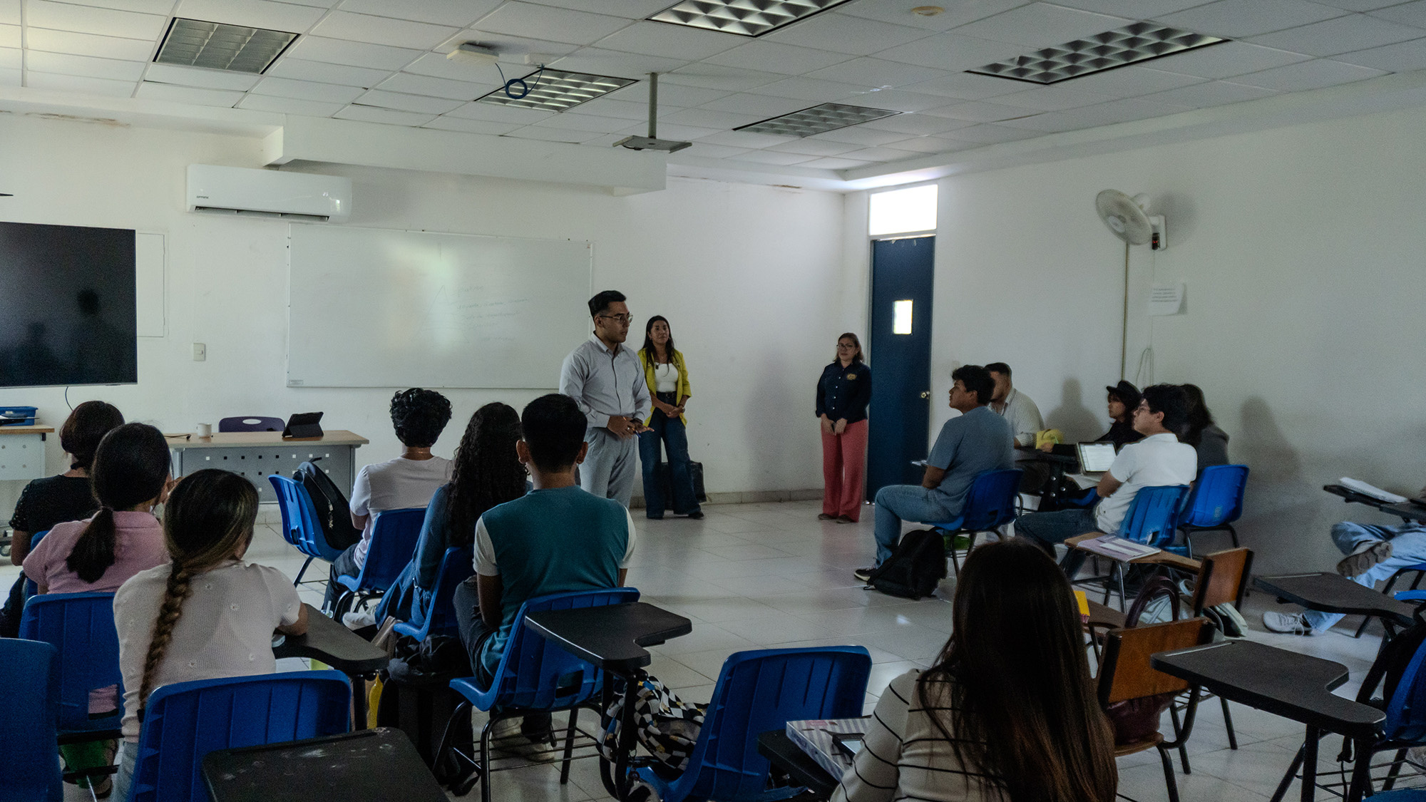 Al menos quince jóvenes sentados en pupitres dentro de un aula, se mantienen atentos a un hombre que habla frente al grupo. Detrás del hombre, hay otras dos mujeres de pie