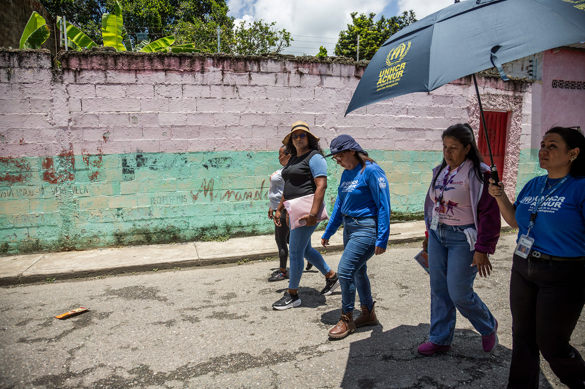 Un grupo de cinco mujeres camina en una calle bajo el sol. Una de ellas sostiene una sombrilla con el logo de ACNUR en amarillo