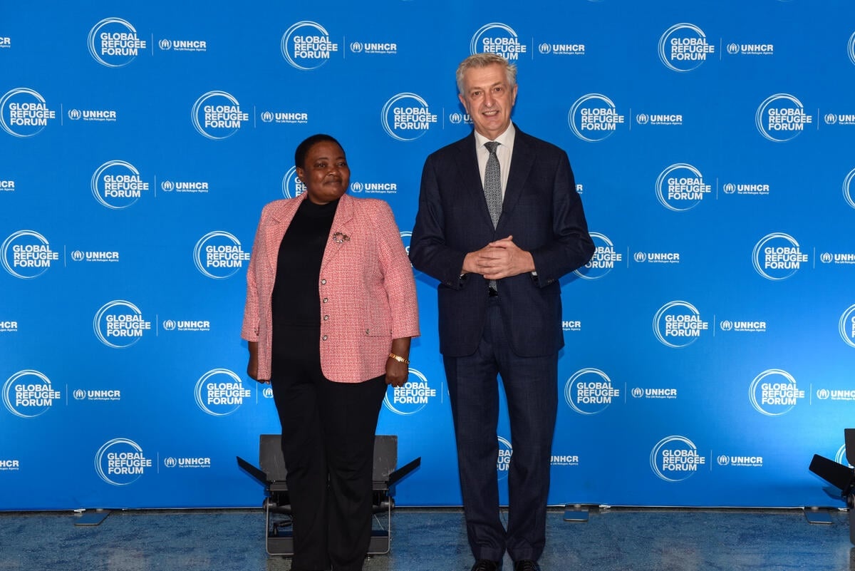 Una mujer y un hombre, ambos con vestimenta formal, de pie frente a un fondo de color azul con logotipos blancos