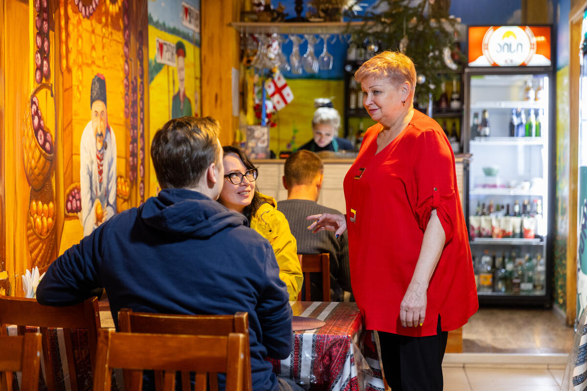 Una mujer de cabello corto que viste de rojo y negro, habla con dos clientes en su cafetería