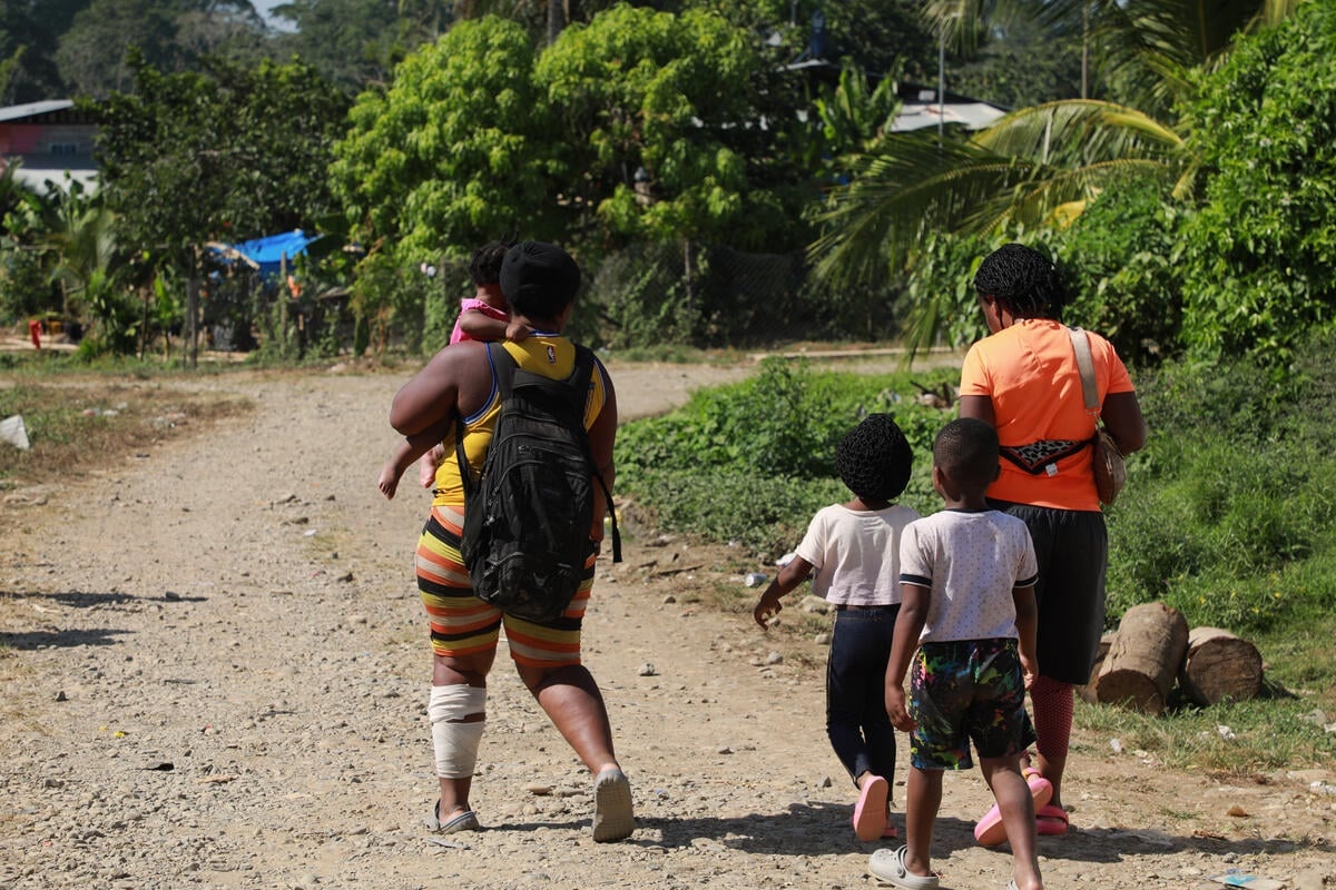 Una mujer y tres niños son fotografiados de espaldas mientras andan por un camino rural. La mujer tiene una pierna vendada y carga en sus brazos a un niño.