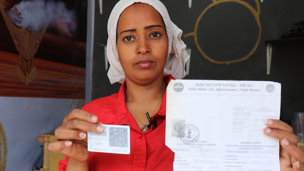 Una mujer con una camisa roja sostiene un documento de identidad en una mano y un papel impreso en la otra.