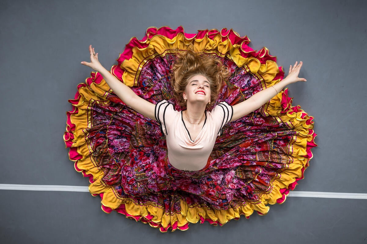 Una mujer posa para una foto tomada desde el aire en una postura de danza que resalta una gran falda colorida que se abre en círculo sobre el piso mientras ella abre sus brazos hacia fuera sonriendo.