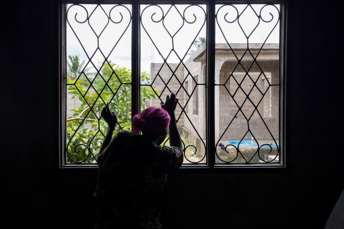 Una mujer de espaldas a la cámara mira por una ventana con las manos en los barrotes de seguridad.