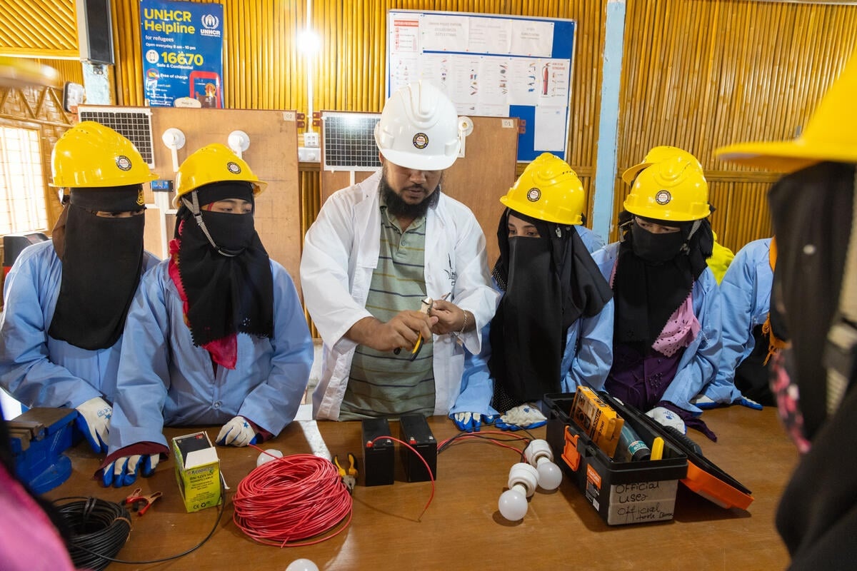 Mujeres con velo negro que les cubre el rostro y la cabeza, y casco amarillo, observan cómo un hombre con bata blanca y casco hace una demostración cortando un cable