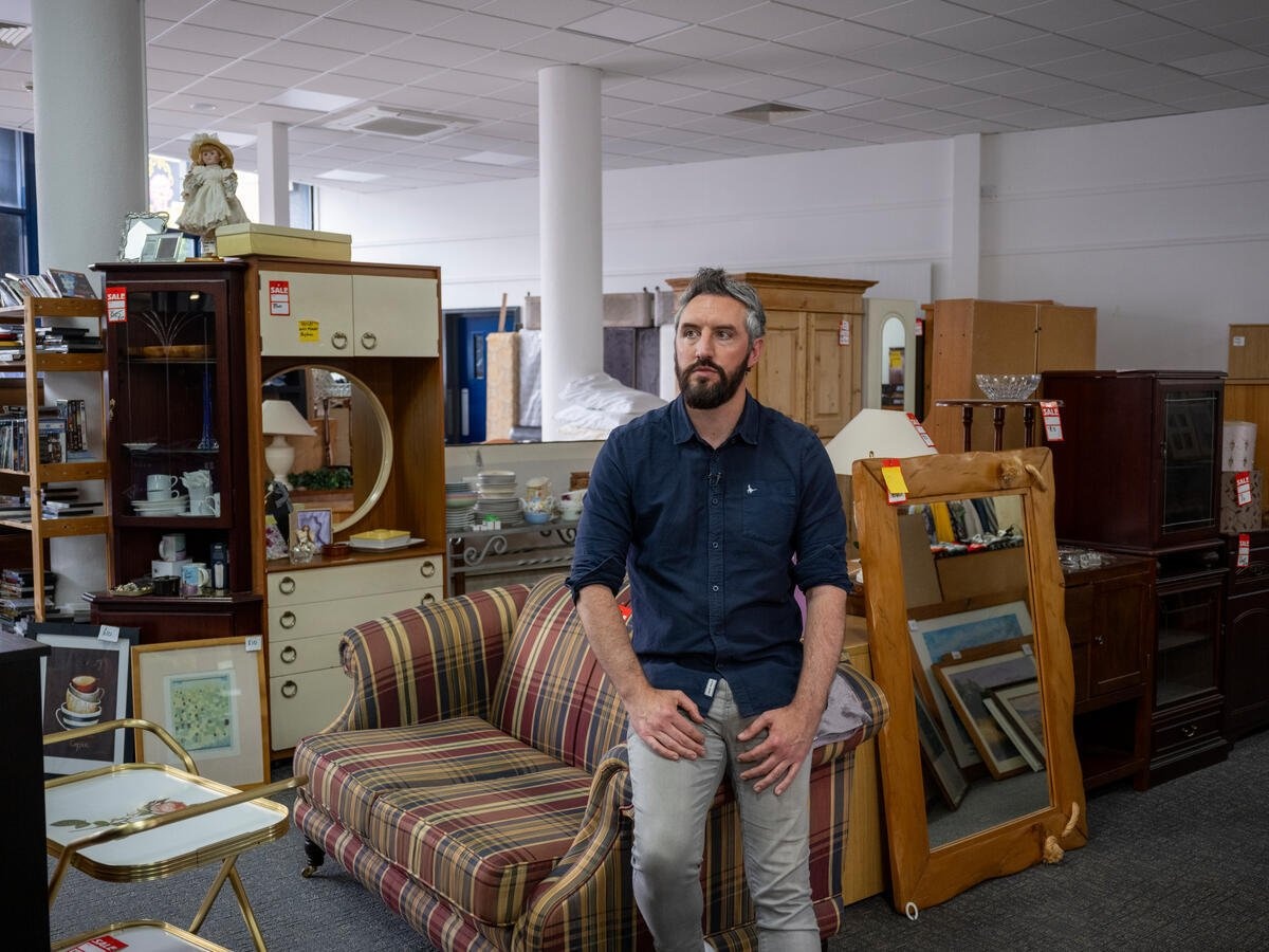 Un hombre con barba vestido con una camisa azul marino y pantalones de mezclilla se apoya en el brazo de un sofá en una tienda de muebles de segunda mano