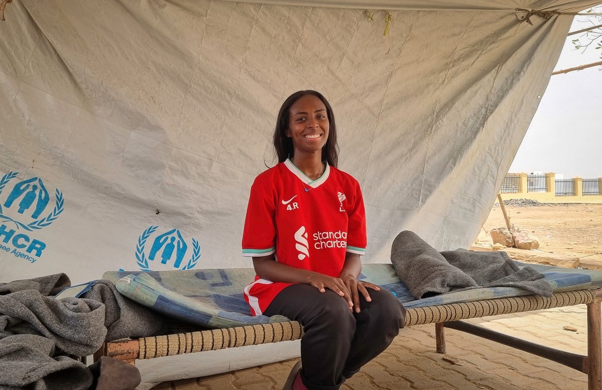 Una joven con una camiseta de fútbol roja en una cama bajo una lona de ACNUR.