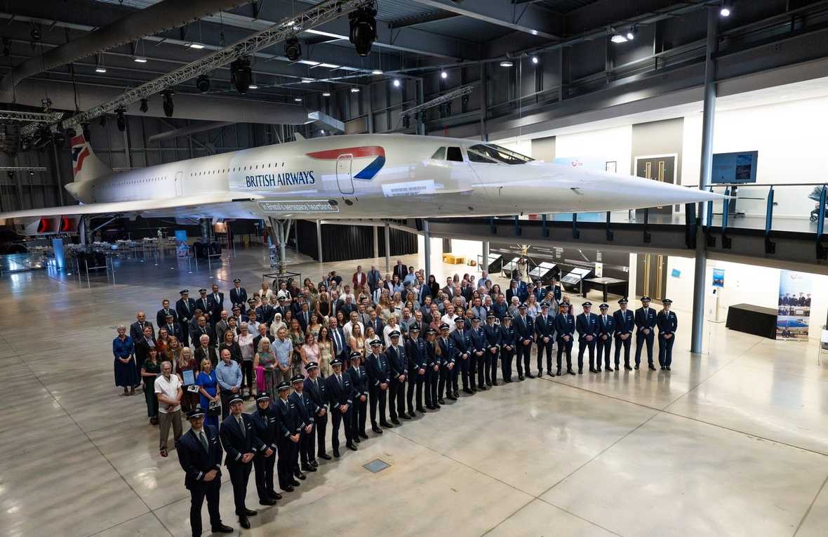 Una fila de pilotos posa delante de un grupo de personas en un hangar con un avión concorde detrás de ellos