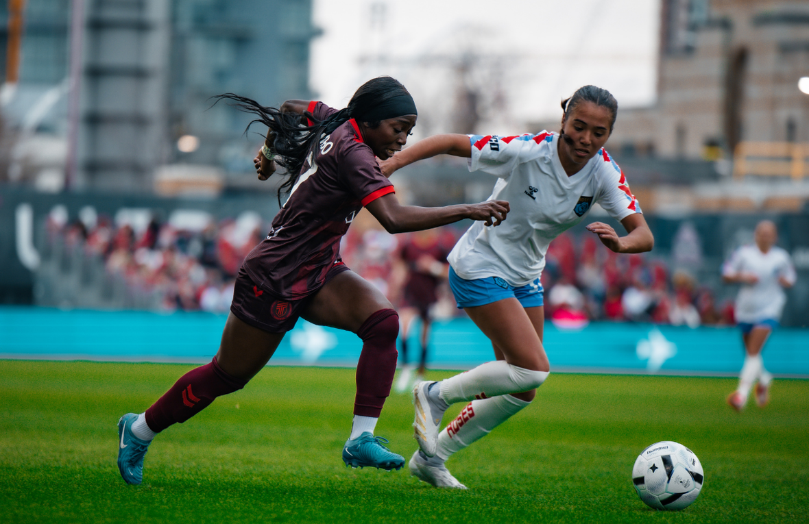 Dos mujeres jóvenes vestidas con uniformes de fútbol de equipos diferentes, corren detrás de un balón en una cancha con pasto verde