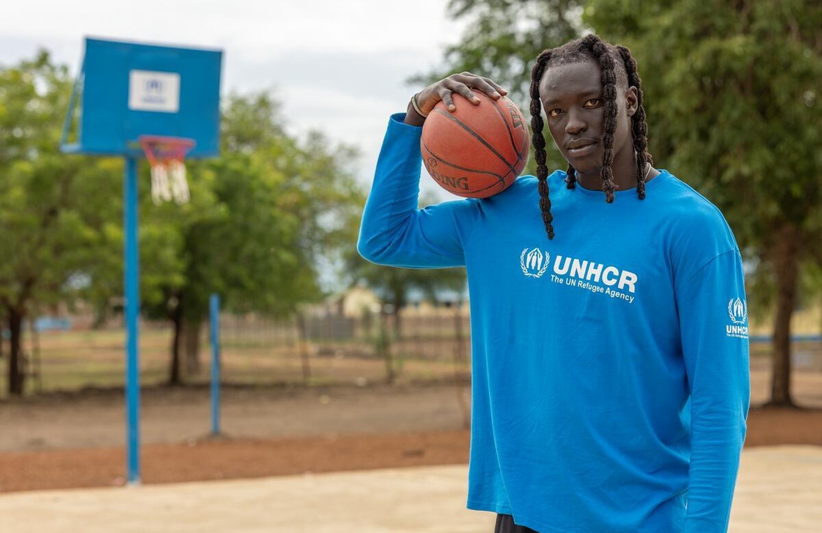 uN hombre con rastas en el cabello que viste una camiseta azul de ACNUR, sostiene una pelota de baloncesto color naranja en una cancha de baloncesto