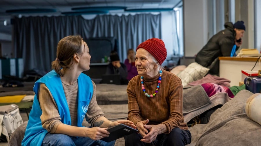 Sentadas sobre una cama en un albergue, una trabajadora de ACNUR charla con una mujer mayor.