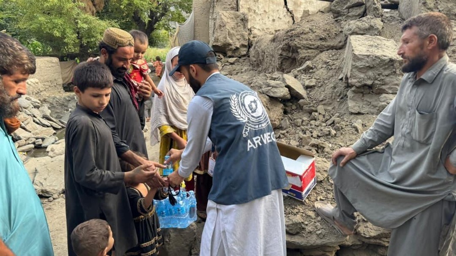 Un hombre con chaleco de visibilidad ARWEO distribuye botellas de agua a un grupo de personas entre los escombros de unos edificios.